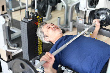 ジムで辛い表情でベンチプレスをする白髪のシニア男性