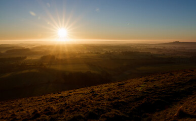 sunburst corona sun flares on the horizon cast a golden glow on the rising mist filled valley of Pewsey Vale from Martinsell Hill, Wiltshire