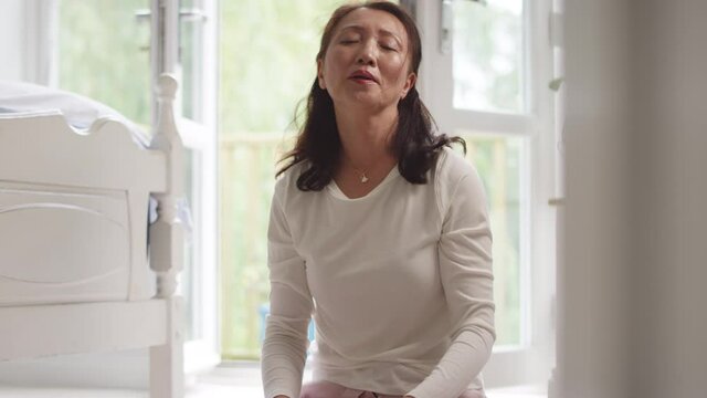 Mature Asian woman in pyjamas sitting on bedroom floor meditating in yoga pose - shot in slow motion
