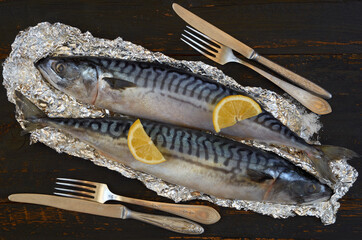 Two atlantic mackerel fishes ready for cooking decorated with spices, salt, lemon slices, knife and fork on the black wooden background. Top view