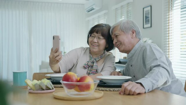 Young senior asia citizen couple hold mobile phone selfie videocall online to family at home apartment morning on dining table in older people with digital technology virtual talk conference concept.