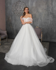 Young beautiful female model wearing long white bridal wedding dress standing and posing in grey interior. Elegant bride