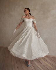 Full length bride portrait. Elegant woman with pony tail in white wedding dress spinning in beige interior. Dynamic shot. Elegant female model standing and posing in ball gown