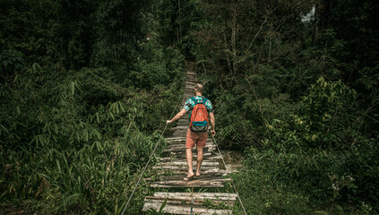 Podróżnik idący przez drewniany most wiszący przez dżunglę. - obrazy, fototapety, plakaty