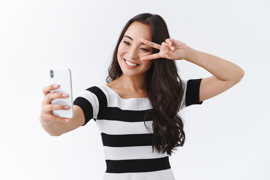 Cheerful, lively good-looking pretty asian woman in casual t-shirt, taking selfie on smartphone, show peace or victory sign near eye, making cute pose to photograph and send pic online