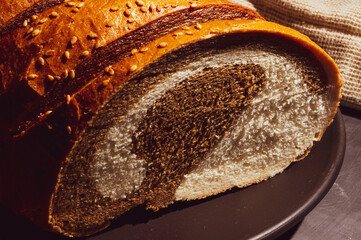 Unusual two-color fresh loaf of bread in a ceramic plate and a woven napkin on a black background, photo in a low key in hard light, close-up.