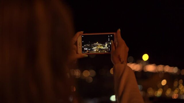  Young woman makes photo of local square illuminated by electric city lights at night holding white smartphone in hands closeup.