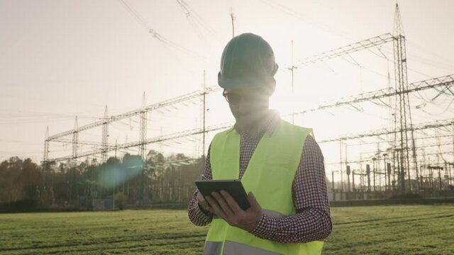 Electrical engineer wearing a helmet and safety vest working with tablet near high voltage electrical lines power station during sunset shot in 4k super slow motion