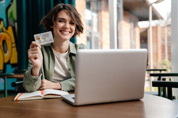 Smiling girl showing credit card while working with laptop in cafe