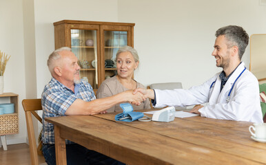 Fototapeta premium Handsome smiling young male doctor and senior man patient shake hands after measuring blood pressure on electronic device. Regular checkup of elderly people by medical digital tonometer at home.
