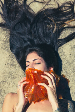 Beautiful tattooed young teenager girl laying on the ground with hair up enjoying sunny day in the street covering face with an orange plastic film. Youth new normal lifestyle portrait concept.