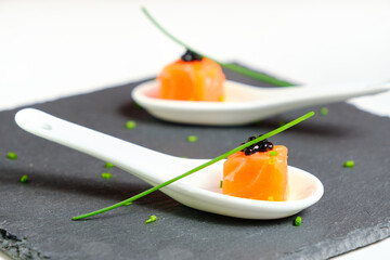 A close-up on a taste of gourmet salmon tartare for catering, with chives and caviar decorating on an elegant white spoon and presented on a black slate