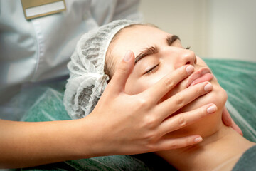 Young caucasian woman receiving facial massage by beautician's hands in spa medical salon