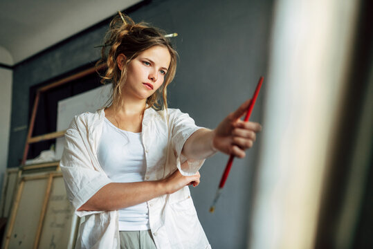 An artist female standing next to the easel with canvas painting something in her art studio. A professional young woman painter searching for an imagination in her workshop.