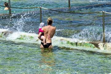 Back view of a woman holidng her baby in a pool along the ocean