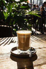 Glass cup with cappuccino on wooden table with hard bright shadows in cafe. Morning sunlight.