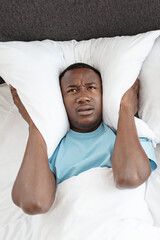 Upset tired young african american man freaking out, covers his ears with pillow so as not to hear...