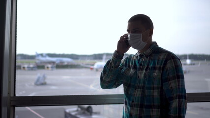 A young man in a medical mask talks on the phone against the background of a window at the airport. Airplanes in the background.