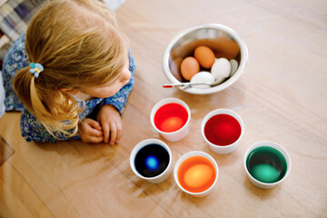Excited little toddler girl coloring eggs for Easter. Cute happy child looking surprised at colorful colored eggs, celebrating holiday with family. From above, unrecognized face.