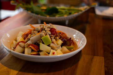 A salad of green apples, red apples, tomatoes, jujube, and peanuts on a white plate on a wooden table in a restaurant. Selective a focus. Take a close-up. Blurred background.