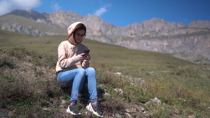 Naklejka na ściany i meble A young woman in a sweatshirt sits with a phone in her hands against a background of mountains.