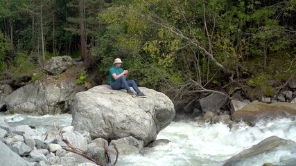 A young man in a Panama hat sits on a stone with a phone in his hands in the mountains by the river.