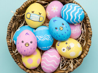 Pandemic holiday. Easter ornament. Quarantine celebration. DIY food decoration. Pastel pink yellow color painted eggs in face mask with chicken bunny design in basket isolated on blue background.