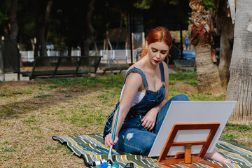 Young caucasian redhead female artist paints a canvas on a small easel with a brush in the park outdoors