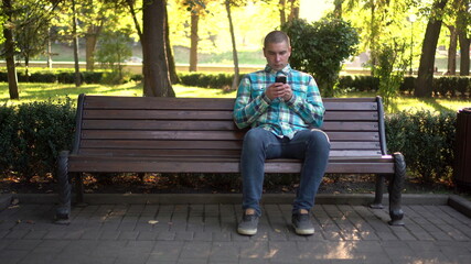 A young man sits on a bench with a phone in her hands. In the background nature.
