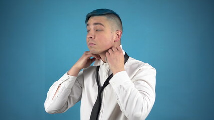 A young man in glasses with blue hair is dressing up a suit. An alternative person fastens buttons and puts a tie on himself on a blue background.