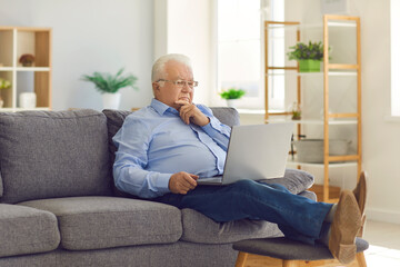 Fototapeta premium Senior man sitting in the room on the couch with a laptop in his hands and thinking.