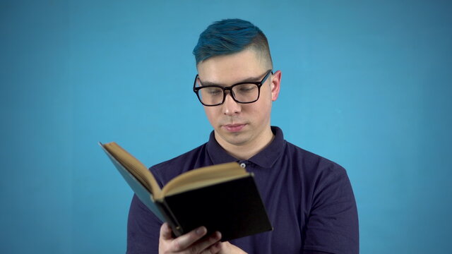 A young man in glasses with blue hair is reading a book. Alternative man with a book in hands on a blue background.