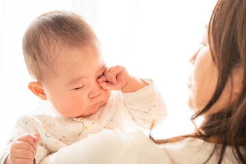 お母さんに抱っこされて眠そうに目をこすっている赤ちゃん