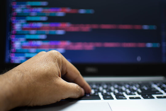 The programmer's hand is writing the source code. Programmers write programs for use with internet business services. The programming code displayed on the laptop screen. Closeup, Blurred background