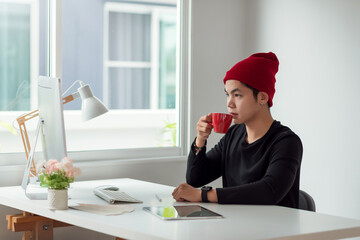 Young designer asian man drinking coffee, analyzing data looking at computer screen at office.