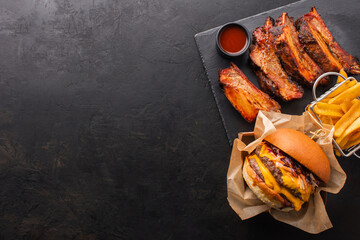 Smoked pork ribs, burger with double cutlets and fries on a black slate board, top view, space for copy.