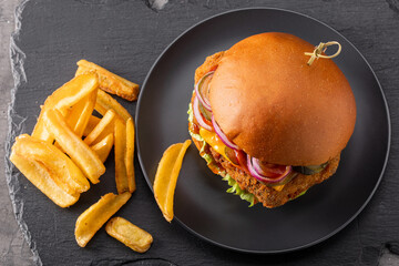 Yummy chicken burger with cheese on a black round plate and fries in side view on a slate background. Fast food set, top view.