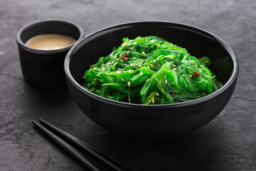 Hiyashi Wakame Chuka healthy seaweed salad with sesame seeds in a black bowl and peanut sauce next to it on a black textured background.
