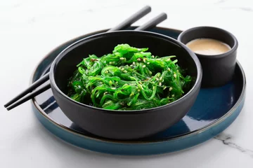 Foto op Canvas Healthy green seaweed salad with sesame seeds in a black bowl and peanut sauce on white stone background, angle view. © Goncharuk film