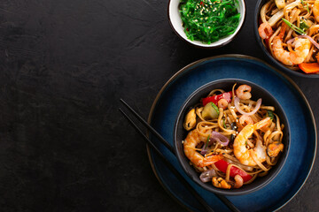 Fried noodles or wok noodles with shrimps, vegetables and mussels in a black bowl and seaweed salad on a black textured background, top view, copy space. Traditional Asian food.