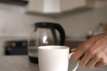 The hand of a young girl takes a white mug from the table against the background of a stylish kitchen.