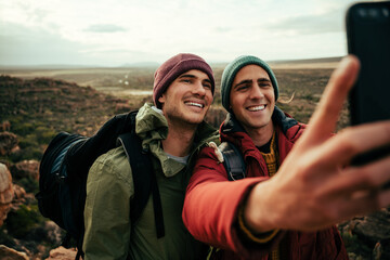 Two caucasian male friends taking selfie with cellular device standing on top of mountain enjoying...