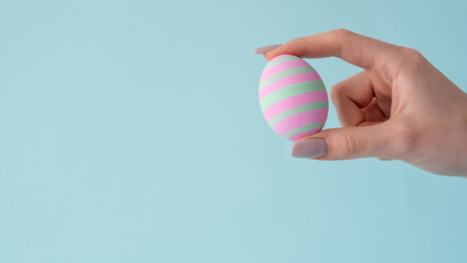 Easter handicraft. DIY decor. Traditional holiday food art. Homemade pink painted egg with mint stripe design in female hand isolated on blue empty space background.