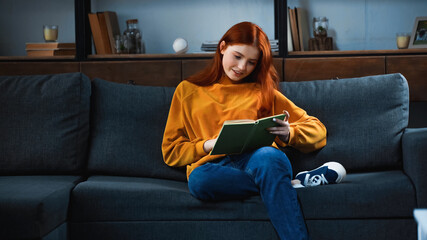 Smiling red haired girl reading book on sofa