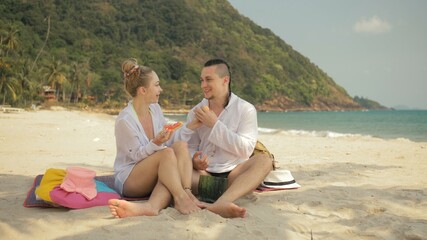 Fototapeta premium The cheerful love couple holding and eating slices of watermelon on tropical sand beach sea. Romantic lovers two people caucasian spend summer weekend in outdoor. Hat, backpack white shirt beachwear.
