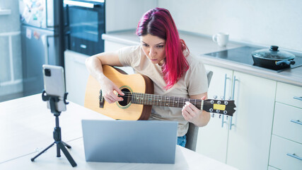 Fototapeta na wymiar A woman sits in the kitchen during a remote acoustic guitar lesson. A girl learns to play the guitar and watches educational videos on a laptop
