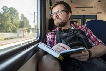 Serious Caucasian man rides a train and looks thoughtfully out the window, in hands he holds a book and a pencil.