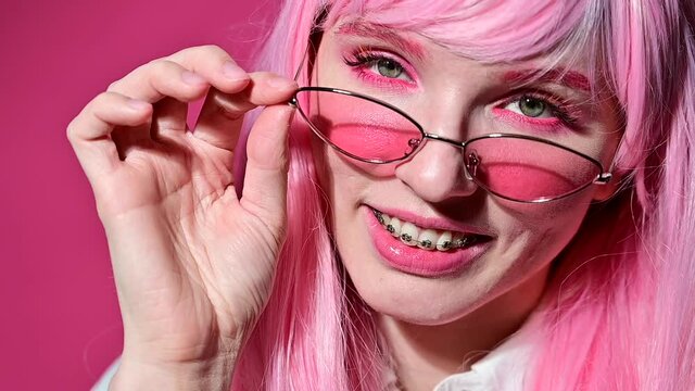 Close-up portrait of a young woman with braces in a pink wig and sunglasses on a pink background.