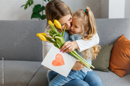 International women's and mother's day. Little cute daughter with bouquet of yellow flowers and postcard congratulates her smiling mom. Happy family holidays at home. Curly girl hugging mommy.