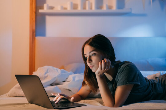 Night online. Bedtime leisure. Internet communication. Relaxed woman using laptop lying in cozy bed in evening home bedroom with warm orange blue light.